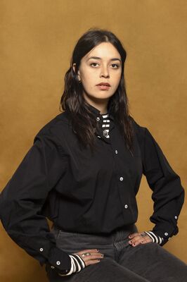 Portretfoto maker Ada Ozdogan van Het Zuidelijk Toneel, poserend met handen in de zij tegen een okergele achtergrond.