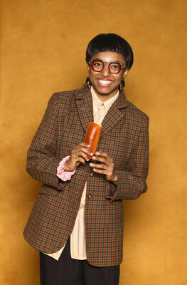 Portretfoto ensemblelid Alicia Boedhoe van Het Zuidelijk Toneel, die poseert met een worstenbroodje tegen een okergele achtergrond.