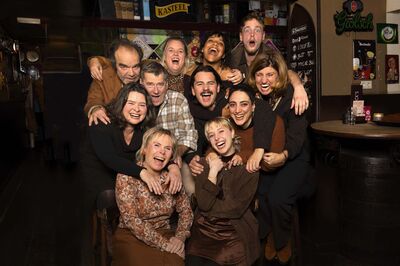 Elf mensen staan als groep lachend op de foto in een bruin cafe