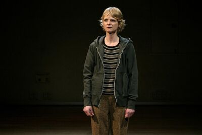 Een jonge actrice in casual kleding, staand op een podium met een donkere achtergrond.