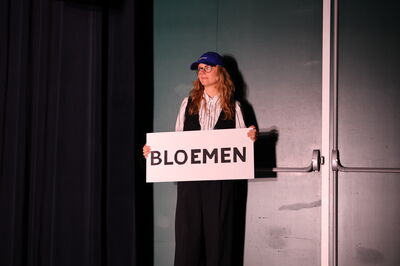 Persoon op een podium met een blauwe pet, houdt een bord omhoog met het woord "BLOEMEN" geschreven.