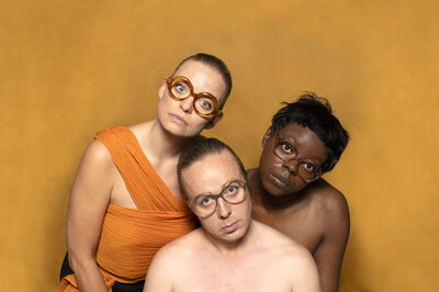 Drie performers poseren dicht bij elkaar tegen een effen okergele achtergrond. Ze dragen grote ronde brillen en kijken met een serieuze, bijna bevreemdende expressie in de camera. Eén persoon draagt een oranje gewaad, de anderen zijn deels ontkleed, wat de intieme en theatrale sfeer versterkt. Het is een campagnebeeld voor de voorstelling Byzantium van Het Zuidelijk Toneel.