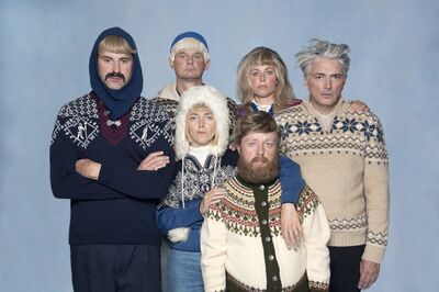 Zes acteurs poseren in winterse kleding met een koude uitdrukking tegen een blauwe achtergrond.