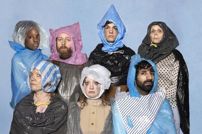Zeven performers staan in twee rijen tegen een lichtblauwe achtergrond, gehuld in capuchons en mantels van gekleurde plastic zakken met serieuze, vermoeide gezichten; het is een campagnebeeld voor de voorstelling De van Het Zuidelijk Toneel in samenwerking met Theater Artemis.