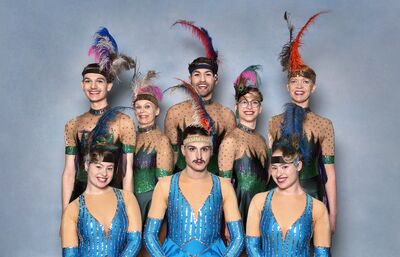 Groep van negen acteurs in kleurrijke kostuums met veren, poseren vrolijk tegen een lichtblauwe achtergrond.