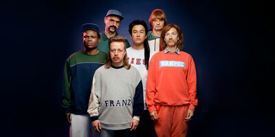 Groep van zes mensen in casual kleding poseren tegen een donkerblauwe achtergrond.