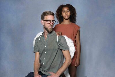 Man met een baard en bril zit op een stoel, terwijl een meisje met krullend haar achter hem staat, tegen een blauwe achtergrond.