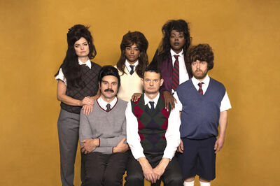 Zes performers in schooluniform-achtige kleding en opvallende pruiken poseren in twee rijen tegen een okergele achtergrond; het is een campagnebeeld voor de voorstelling Not Quichot van Het Zuidelijk Toneel.
