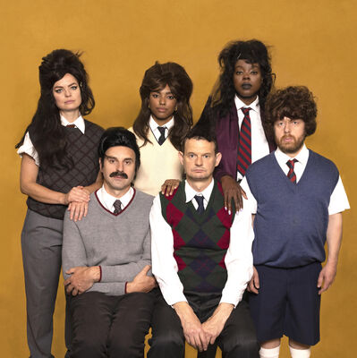 Zes performers in schooluniform-achtige kleding en opvallende pruiken poseren in twee rijen tegen een okergele achtergrond; het is een campagnebeeld voor de voorstelling Not Quichot van Het Zuidelijk Toneel.