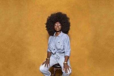 Julia Ghysels zit, met een grote afro en een denim outfit, op een kruk tegen een okergele achtergrond en kijkt rustig in de camera; het is een campagnebeeld voor de voorstelling Saved Game van Het Zuidelijk Toneel.