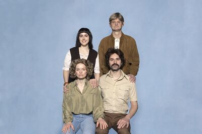 Groep van vier acteurs poserende voor een blauwe achtergrond, met verschillende kledingstijlen en lengtes van haar.