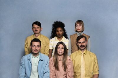 Zes acteurs zitten en staan voor een blauwe achtergrond, gekleed in verschillende pastelkleuren.
