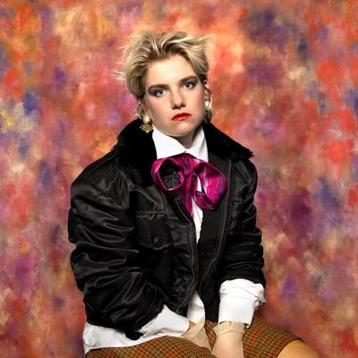 Performer Roos Bottinga poseert verveeld voor een kleurrijke achtergrond. Zij draagt een opvallende shawl, een zwart colbertje en knalrode lippenstift. Dit is het campagnebeeld voor de educatievoorstelling 'Bek Dicht' van Het Zuidelijk Toneel.