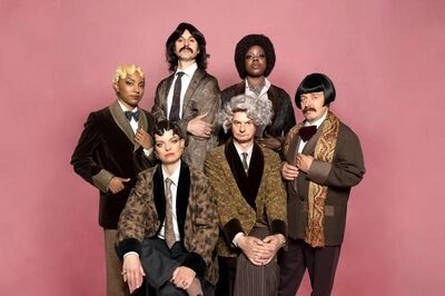 Zes performers in kostuums met verschillende kapsels poseren tegen een roze achtergrond.