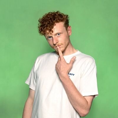 Performer Teun Donders met krullend haar in T-shirt staat tegen een groene achtergrond en maakt een gebaar met zijn hand bij zijn gezicht.