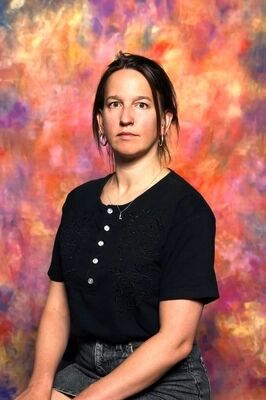 Portretfoto regisseur Lara van Hoof in zwarte top met knopen, zittend voor een kleurrijke, abstracte achtergrond.