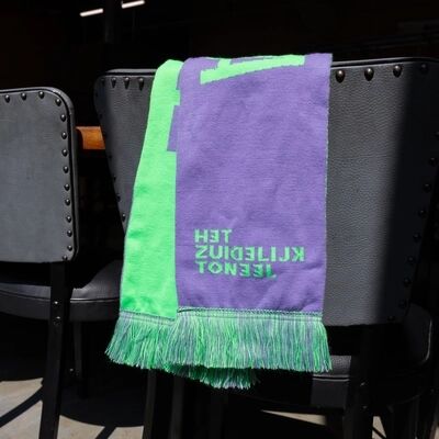 Een kleurrijke sjaal van Het Zuidelijk Toneel in paars en groen met franjes, hangt over een donkere theaterstoel.