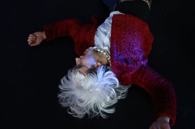 Een persoon met een witte pruik ligt op de grond, gekleed in een rode trui en een witte blouse.