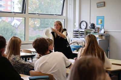 Een workshop-docente van Het Zuidelijk Toneel staat voor een groep middelbare scholieren in een klaslokaal. Ze praat en gebaart enthousiast richting de leerlingen, die aan tafels zitten en aandachtig luisteren. Op de achtergrond staan een kast met boeken, een klok en grote ramen die uitkijken op een groen buitenterrein.