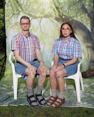 Man en vrouw zijn suf gekleed, kijken streng en zitten op witte plastic tuinstoelen voor een doek waarop een groen park staat.