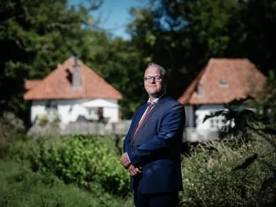 Joris van Bengevoord, een man in een donkerblauw pak met bril en rode stropdas, staat buiten in een groene, bosrijke omgeving. Achter hem staan twee witte huizen met oranje pannendaken. Hij kijkt recht in de camera en poseert met zijn handen voor zich gevouwen.