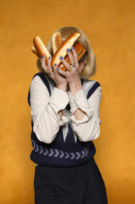 Portretfoto ensemblelid Keja Klaasje Kwestro van Het Zuidelijk Toneel, die poseert met worstenbroodjes tegen een okergele achtergrond.
