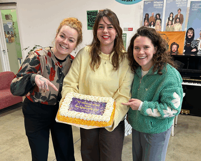 Drie vrouwen houden een slagroomtaart met confetti vast. Op de taart staat geschreven 'Het Zuidelijk Toneel blijft Het Zuidelijk Toneel'.