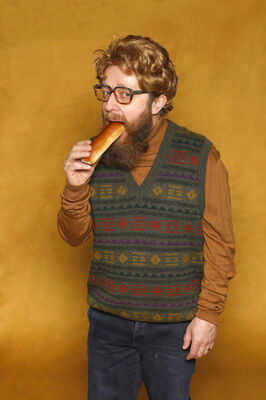 Portretfoto ensemblelid Louis van der Waal van Het Zuidelijk Toneel, die poseert met een worstenbroodje tegen een okergele achtergrond.