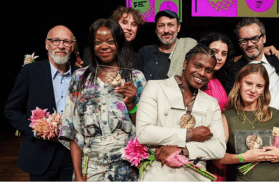 Een groep performers en makers staat bijeen op een podium tijdens een prijsuitreiking. Verschillende personen houden een medaille of bloemen vast en kijken glimlachend in de camera. De sfeer is feestelijk, met kleurrijke bloemen en een achtergrond met felgekleurde grafische panelen.