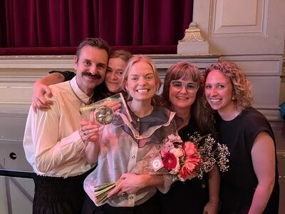 Vijf personen poseren voor een theatrale setting, lachend met een prijs (Theo d'Or) in handen van de persoon in het midden.