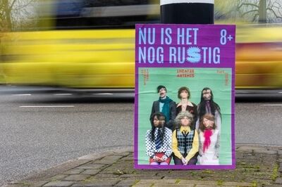 Poster van voorstelling Nu is het nog Rustig staat geplakt op posterbord op straat en er rijdt een gele bus snel voorbij.