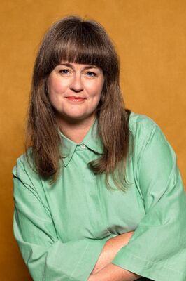 Portretfoto maker Sarah Moeremans van Het Zuidelijk Toneel, poserend met armen over elkaar tegen een okergele achtergrond.