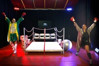 Twee mensen in boksoutfits staan enthousiast in een boksring, met een podium en sportattributen op de achtergrond.