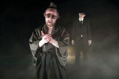 Louis van der Waal spreekt met bebloede handen naar het publiek terwijl een andere performer in een donker pak met zonnebril stil op de achtergrond staat.