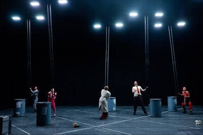 Zes performers staan verspreid over een toneel, ieder naast een grote blauwe ton en een hoge, verticale metalen constructie die naar de felle theaterlampen boven hen reikt. Ze kijken omhoog of trekken aan de constructies, terwijl bloemen op de vloer liggen. De scène heeft een dramatische, experimentele uitstraling in een verder donkere ruimte.