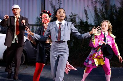 Vier performers spelen een dynamische scène waarin één persoon in een grijs pak naar voren stapt terwijl de anderen achter haar reageren.