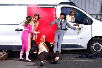 Vijf performers spelen een chaotische scène rond een bestelbus waar rode rook uit de open schuifdeur komt.