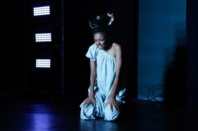 Alicia Boedhoe knielt in blauw licht op het toneel, gekleed in een asymmetrische jurk en met takken verwerkt in haar haar.