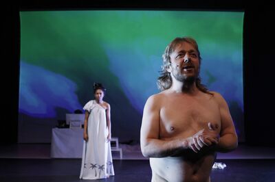 Op de voorgrond staat Louis van der Waal met een ontbloot bovenlijst te spreken terwijl Alicia Boedhoe in een lichte jurk achter hem voor een kleurrijke projectie staat.