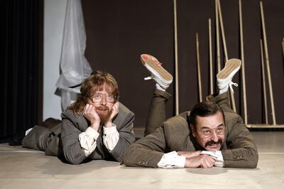 Louis van der Waal en Joep van der Geest liggen voorover op de grond in kostuums en kijken op speelse wijze voor zich uit.