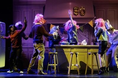 Een groep mensen met kleurrijke pruiken danst in een keuken tijdens een feestelijke voorstelling.