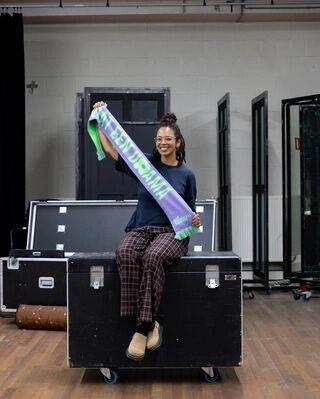 Vrouw poseert op theaterboxen met groen-paarse supporterssjaal van Het Zuidelijk Toneel en houdt deze schuin omhoog.