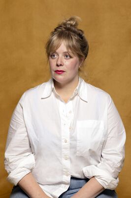 Portretfoto maker Suze Milius van Het Zuidelijk Toneel, poserend tegen een okergele achtergrond.