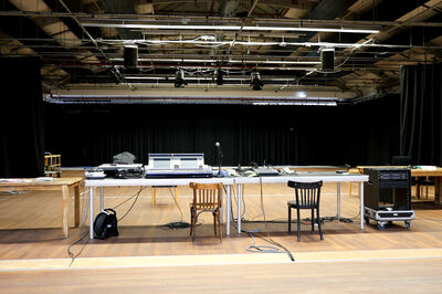 Een lege oefenruimte met een mengtafel, enkele stoelen en zwarte gordijnen op de achtergrond.